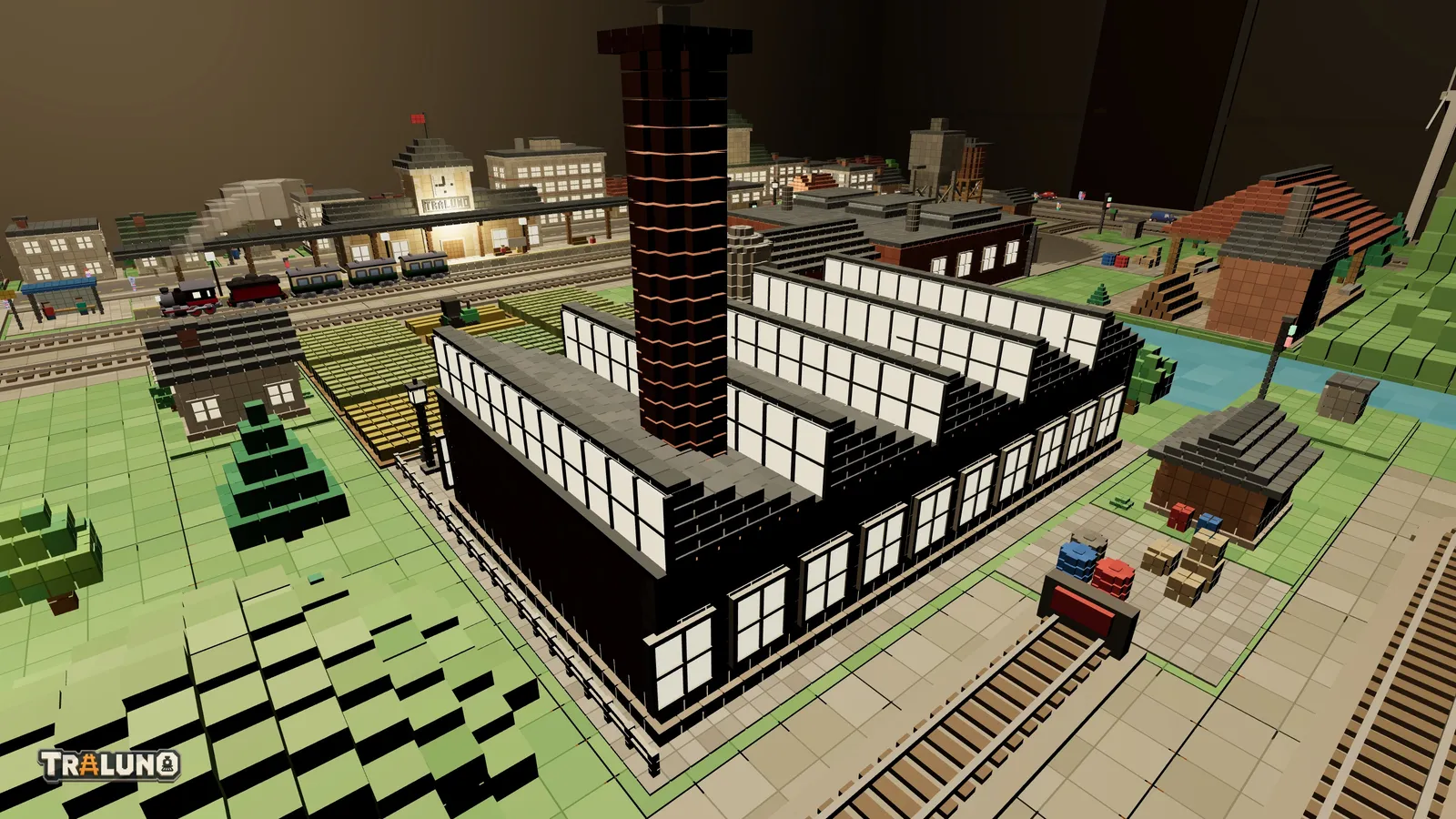 The Brick Factory at night seen from behind: the glazed faces of its four-bay sawtooth roof glowing white, a row of lit tall windows down the long side, the brick chimney rising from the middle of the roof, and the station and town lit beyond.